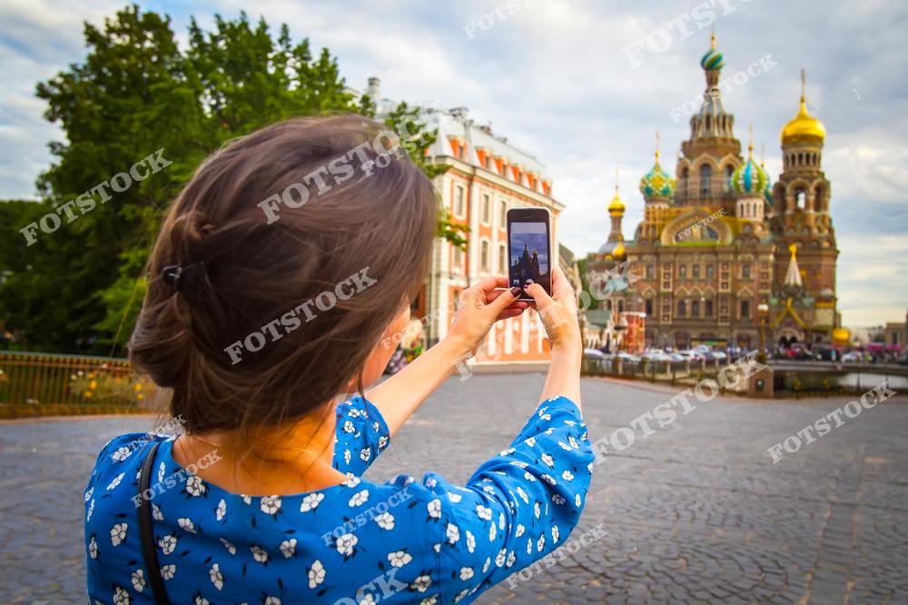 На снимке девушка делает фотографию собора Спаса-на-Крови в Санкт-Петербурге с помощью смартфона. Фоном выступает величественное здание собора с его характерными куполами и орнаментами. Зеленые насаждения и мощеная мостовая добавляют атмосферности и подчеркивают исторический облик города. Это место — популярная туристическая достопримечательность и символ культурного наследия Петербурга.